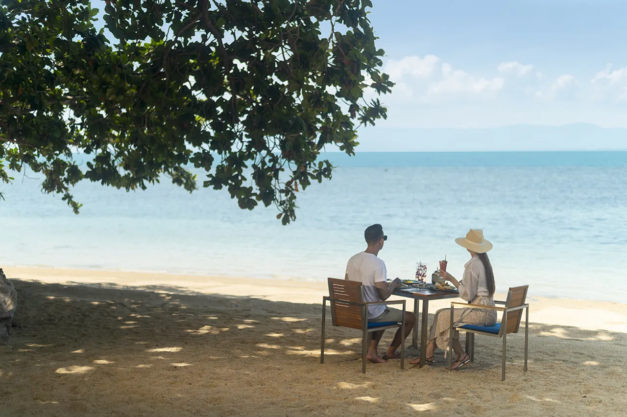 A couple enjoying a beachfront lunch at Explorar Koh Phangan, with gourmet dishes and stunning ocean views enhancing their perfect tropical escape.